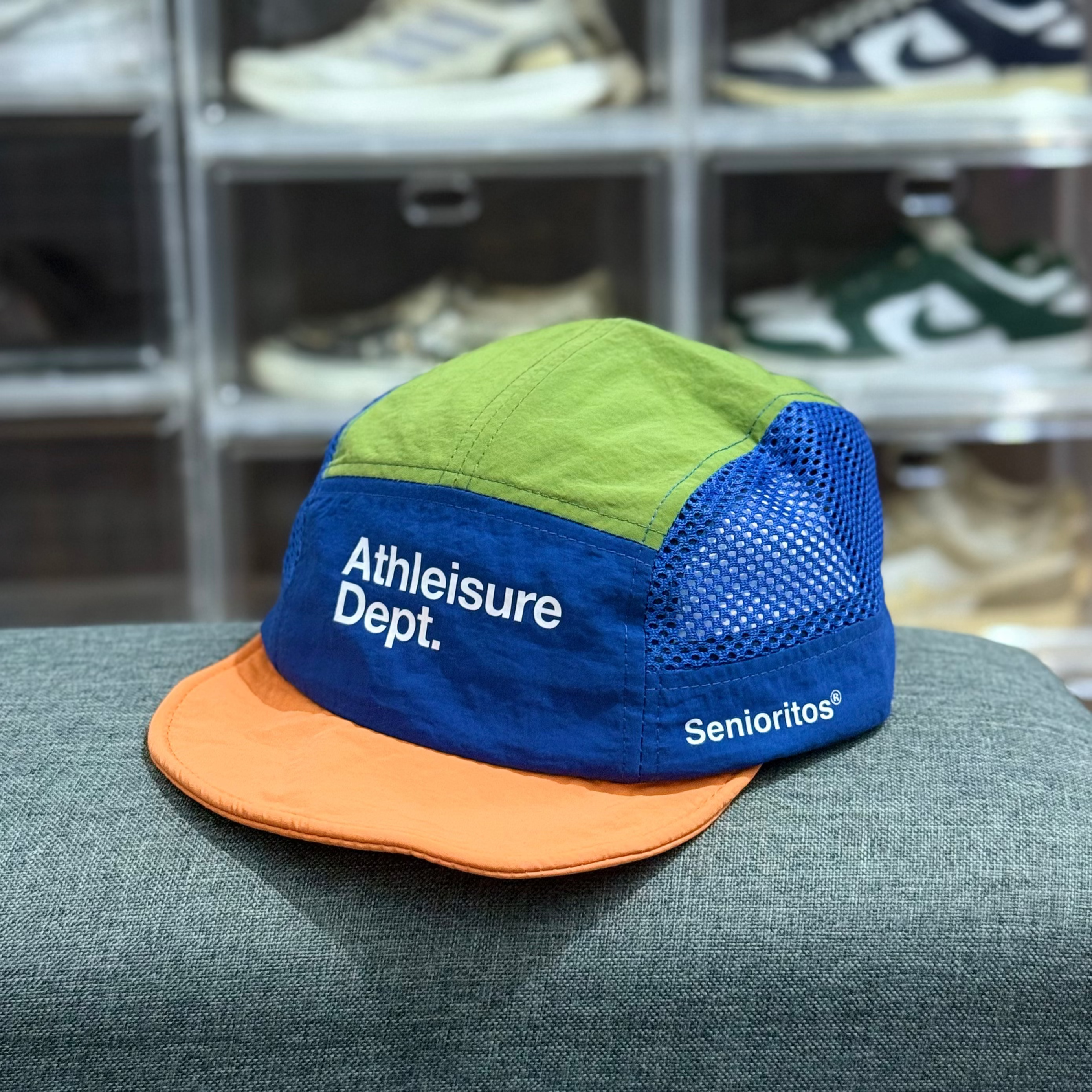Colorful cap with 'Athleisure Dept.' and 'Senioritos' text on a gray surface with shoes in the background